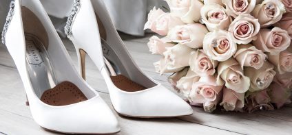 Weiße Stöckelschuhe mit Glitzerapplikationen, daneben ein Brautstrauß aus hell-rosa Rosen und im Hintergrund ist ein weißes Kleid zu erkennen.