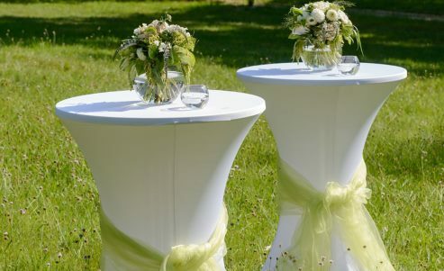 Stehtische im Freien auf einer grünen Wiese mit weißem Bezug und Blumenvasen in der Tischmitte.