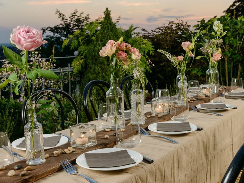 Eine lange Tafel mit Tellern, Servietten und Besteck sowie Dekoration mit rosa Rosen.