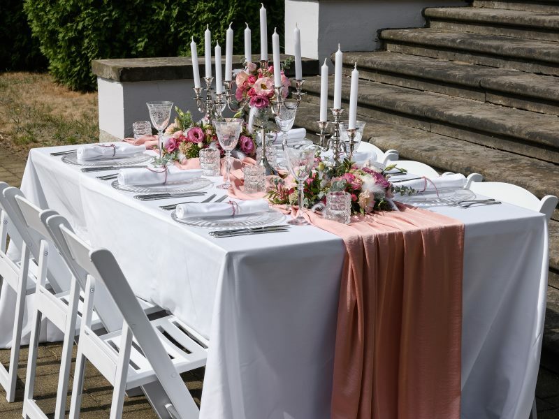 Eine lange gedeckte Tafel mit weißer Tischdecke, weißen Stühlen, Kerzenständern mit weißen Kerzen sowie Dekoration in rosa.