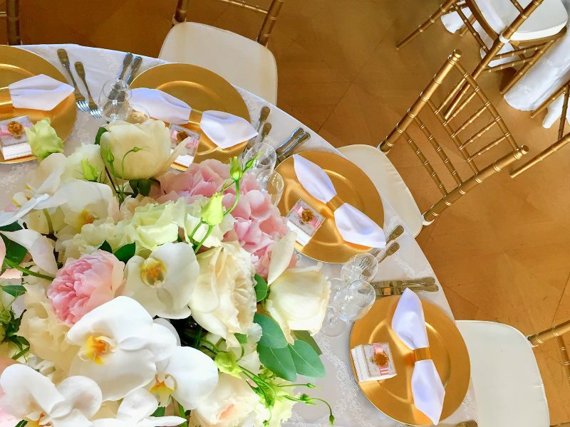 Ein runder Tisch mit goldenen Stühlen, Tellern und Serviettenhaltern. In der Mitte befindet sich ein großes Blumenarrangement mit Orchideen.