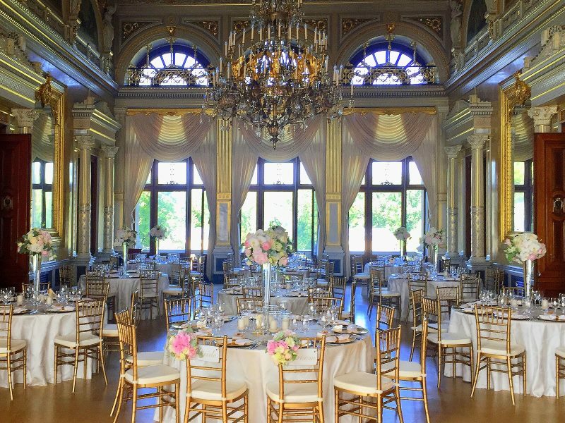 Ein großer Raum mit Verzierungen, einem großen Kronleuchter in der Mitte und neun runden Tischen. Die Dekoration, Stühle und Geschirr sind gold und teilweise mit Orchideen geschmückt.