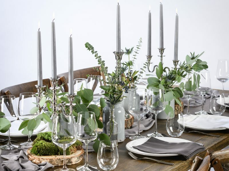 Ein gedeckter Tisch mit weißen Tellern, grauen Servietten, Gläsern und drei Kerzenständer in der Mitte. Um diese herum befinden sich diverse grüne Gestecke in Ton-Vasen.