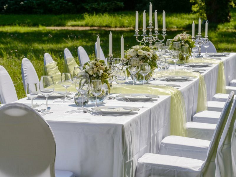 Eine lange gedeckte Tafel mit weißer Tischdecke, weißen Stuhlhussen, Kerzenständern mit weißen Kerzen sowie Dekoration in grün.