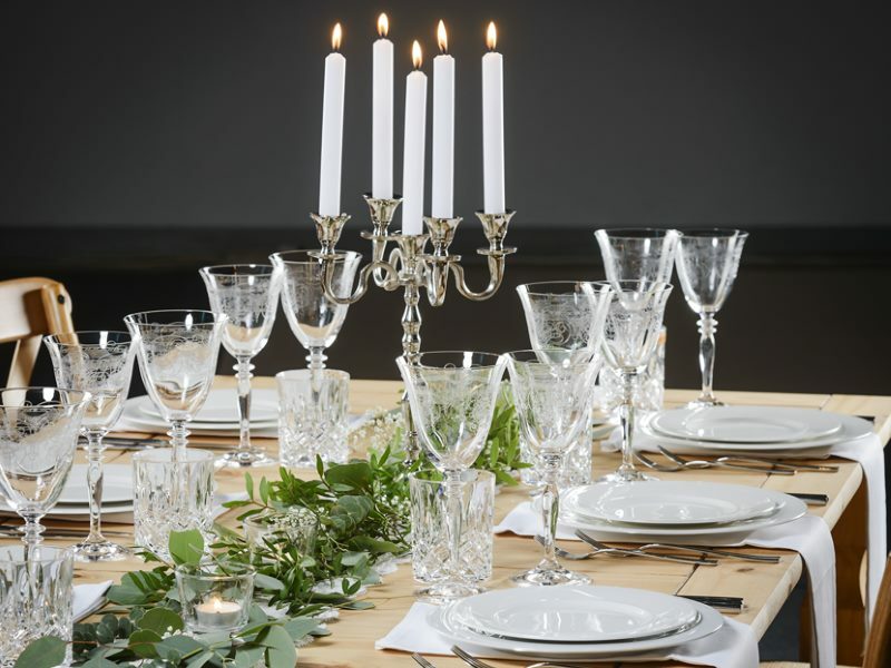 Ein gedeckter Tisch mit weißen Tellern, Gläsern und einem Kerzenhalter in der Mitte. Um diesen herum ist ein langes Gesteck aus grünen Blättern, das entlang des ganzen Tisches geht.