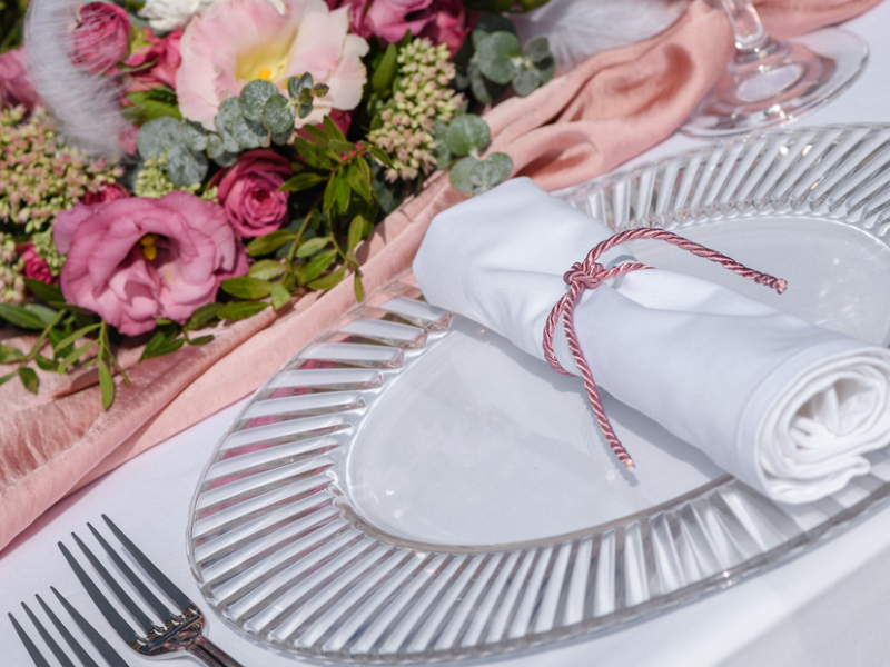 Ein Platzteller mit Serviette, um die ein rosanes Band gebunden wurde. Im Hintergrund sieht man ein Blumengesteck mit rosa Blüten.