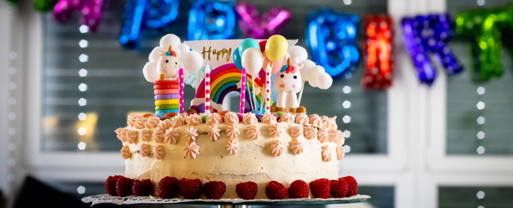 Eine große Torte mit Verzierungen, Himbeeren, Kerzen und einer Karte. Im Hintergrund hängt ein bunter Happy Birthday Schriftzug über drei Fenstern.
