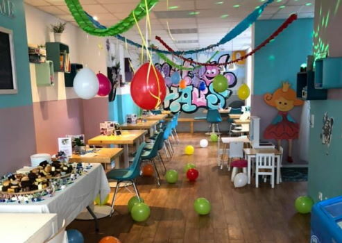 Das Innere vom Mutzelhaus Dresden: Viele Stühle und Tische, auf einem Tisch sind leckere Speisen. Des Weiteren gibt es bunte Luftballons, Girlanden und Luftschlangen zu sehen.