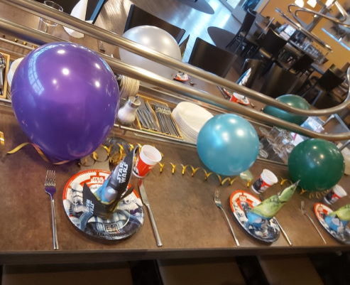 Tischdekoration zum Kindergeburtstag: bunte Luftballons, Luftschlangen sowie Teller und Servietten mit „Star Wars“ Motiv. In der Mitte der Tafel ist eine Schiene, wo das Essen serviert wird.