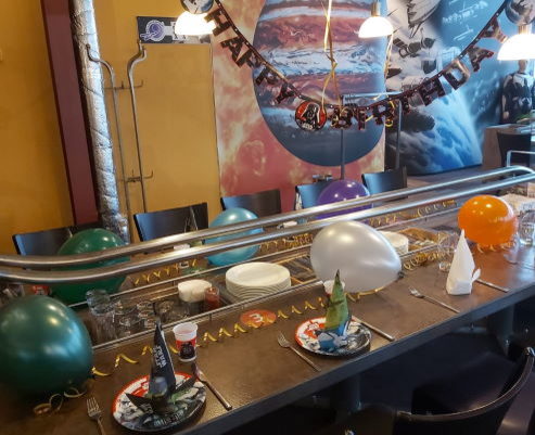 Tischdekoration zum Kindergeburtstag: bunte Luftballons, Luftschlangen, eine Geburtstagsgirlande sowie Teller und Servietten mit „Star Wars“ Motiv. In der Mitte der Tafel ist eine Schiene, wo das Essen serviert wird.