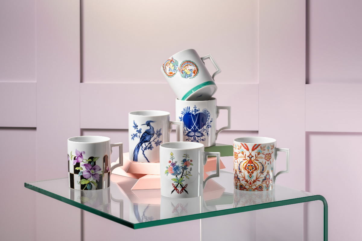 Ausgewählte Porzellanstücke von MEISSEN, präsentiert auf und an den pinken Verpackungen sowie auf einem kleinen Tisch. Rechts steht eine große blau-weiße Vase und zwischen den Stücken befinden sich lange Grashalme.
