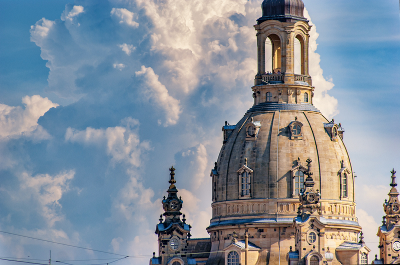 Die impulsante Dresdner Frauenkirche mit einer Nahaufnahme der Kuppel, im Hintergrund blauer Himmel und eine aufsteigende Wolkenfront.